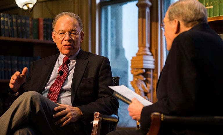 Hugh Price sits with legs crossed on a riser talking to Peter Edelman during an event in Riggs Library.