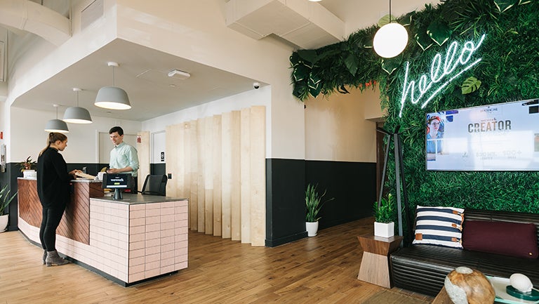 man assisting woman at front desk at WeWork White House space with plant-covered wall, the word hello in lights, sofa and pillow