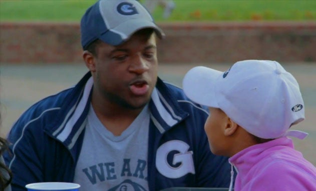 Justin Pinn, squatting and wearing a Georgetown jacket, T-shirt and hat talks with child wearing baseball cap