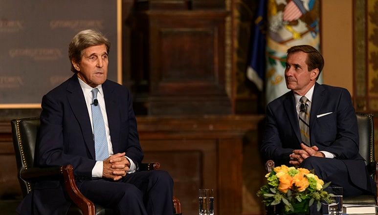 John Kerry sitting onstage with hands folded in lap next to John Kirby