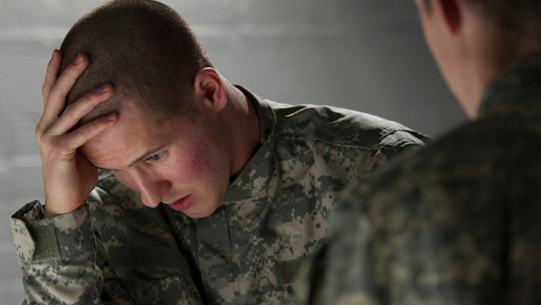 Soldier in uniform, sitting with head in hand with other soldier standing over him