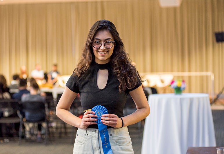 Maddy Rice stands holding blue ribbon with chairs tables and stage curtain behind her