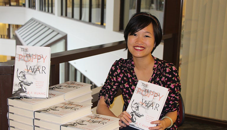 Rebecca Kuang holds up a copy of her book The Poppy War surrounded by numerous other copies 