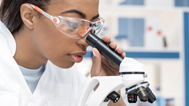 woman wearing goggles and lab coat looks into microscope