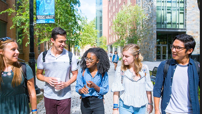 Five students walk outside on campus near Pedro Arrupe, S.J. Hall near a banner reading "Educating the Whole Person."