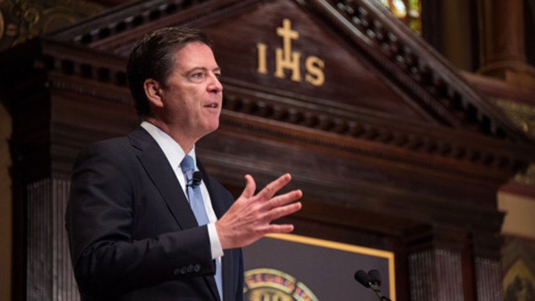 FBI Director James Comey standing and speaking in Gaston Hall with the Jesuit IHS sign behind him