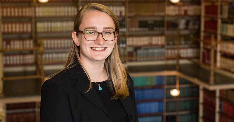 Annee Lyons smiles into the camera wearing glasses with a wall of library books on shelves in the background.