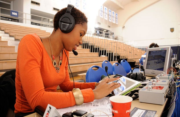 Monica McNutt sits behind a table with papers and a TV set with a headset on while taking notes in McDonough Arena