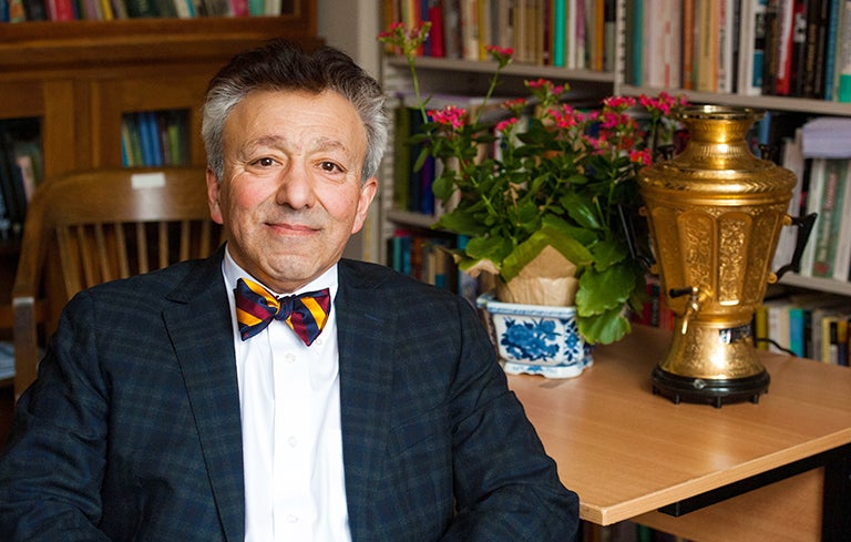 Fathali Moghaddam sits at a table with a gold vase and pink flowers in front of shelves of books. 