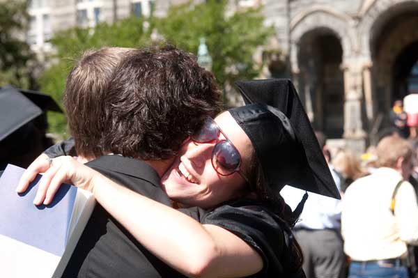 A graduate hugs someone while smiling in sunglasses with Healy Hall in the background
