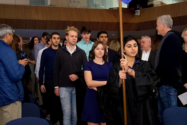 A student leads the way holding up a school banner as other students follow her inside the auditorium as family members look on.