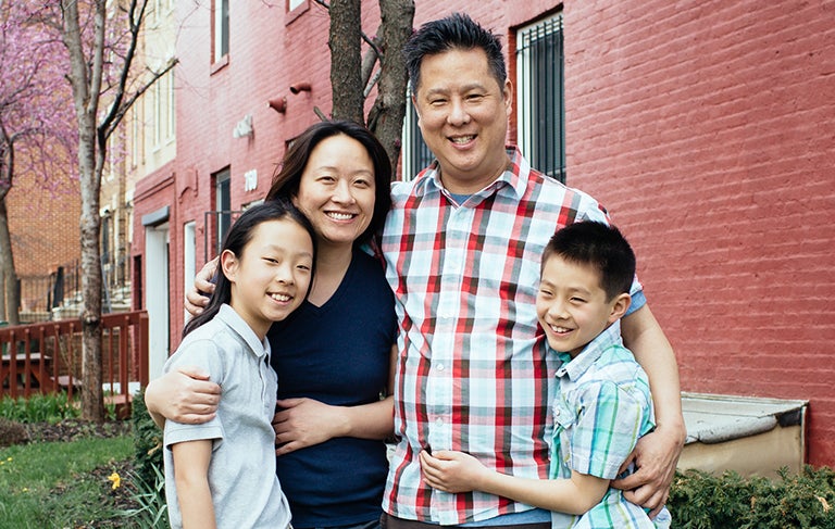 Steve Park stands with his wife and children outside in front of the side of a rowhome.