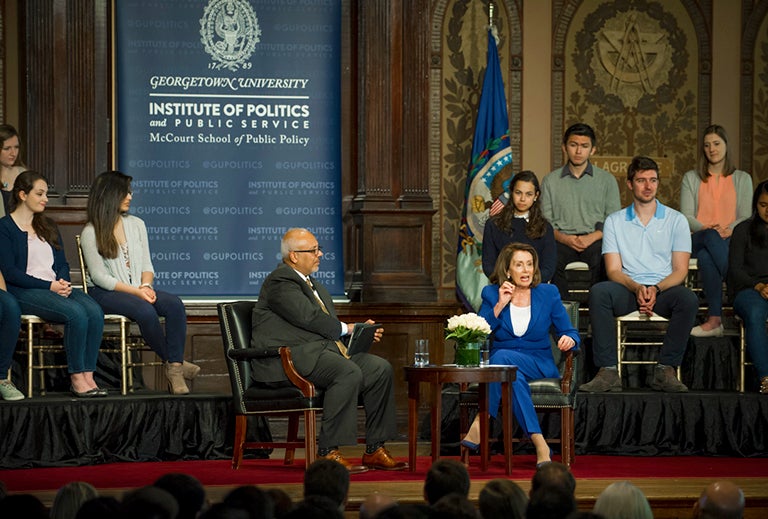 Mo Elleithee sits on stage with Nancy Pelosi with students sitting on risers in the background.