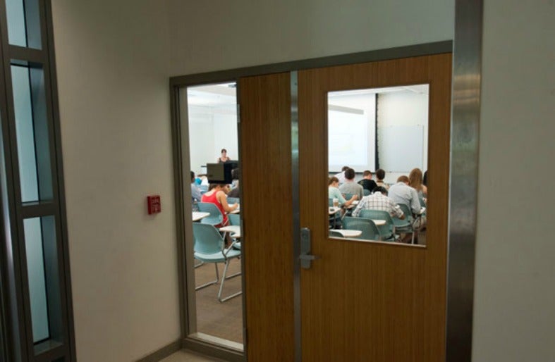 A window on a door shows the inside of a classroom with students and a professor lecturing