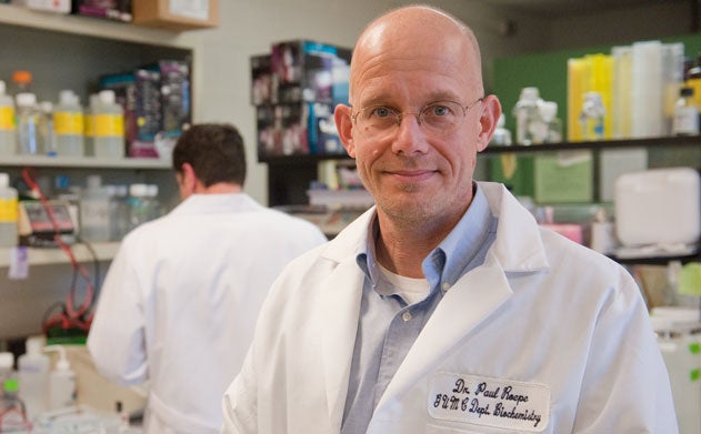 Paul Roepe, wears a lab coat, and researchers work in the lab behind him