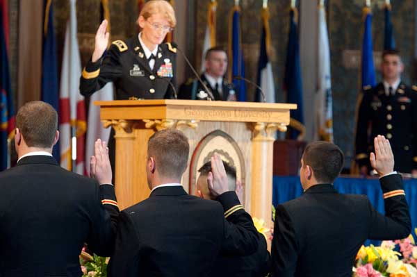 Mary Legere raises her right hand behind a podium as members of the audience in military uniform do the same