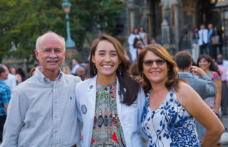 Hailey Roumimper wears her medical school white coat as she poses with her parents in front of Healy Hall.