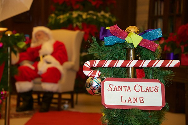 A man dressed as Santa sits in the background in a chair while a sign for Santa Claus Lane is focused on