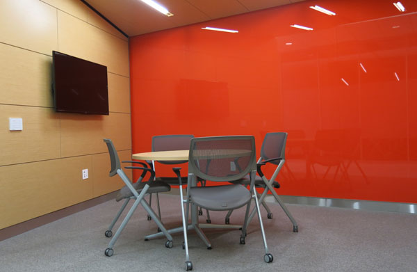 An SCS breakout room has bold orange-red accent wall, wall-mounted LCD sceen and modern meeting table and chairs