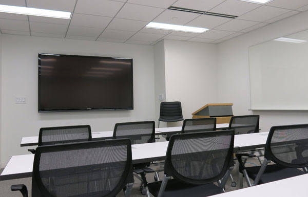 A bright white classroom features wall-mounted LCD screen and wheeled black office chairs