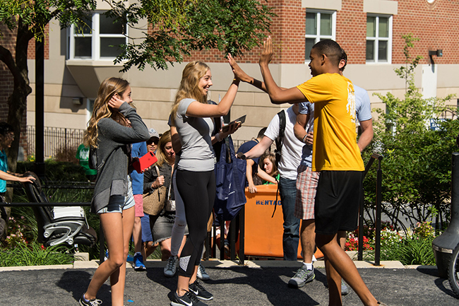 a new student orientation team member welcomes a new student on campus with a high-five as another new student looks on.