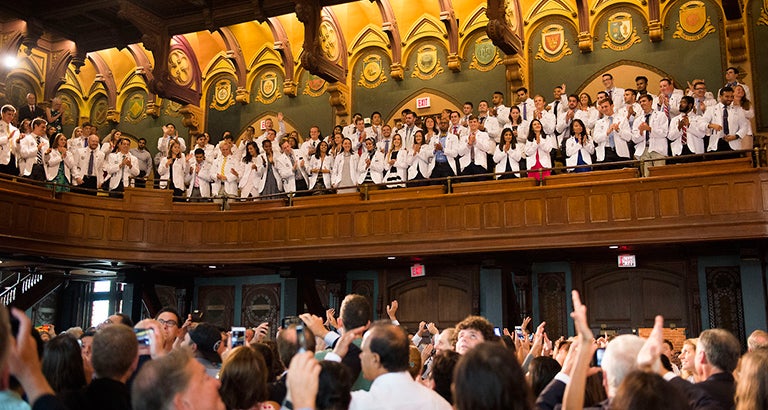 Medical students stand in the balcony clapping as they look down at the audience looking up to them and clapping for them